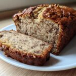 A loaf of moist sugar free banana bread with one thick slice cut and resting beside it on a white plate.