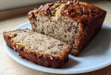 A loaf of moist sugar free banana bread with one thick slice cut and resting beside it on a white plate.