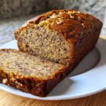Close-up of a loaf of moist sugar free banana bread, with one slice cut and resting on a white plate.