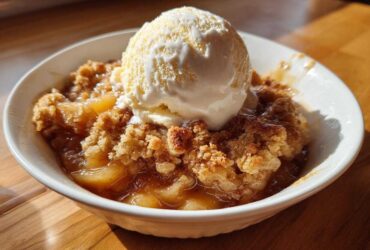A close-up of a warm apple cobbler recipe served in a white bowl, topped with a melting scoop of vanilla ice cream.