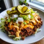 A close-up of a healthy cheeseburger bowl featuring seasoned ground beef, melted cheese, shredded lettuce, onions, and pickles.
