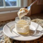 A spoonful of thick, creamy homemade mayonnaise is being lifted from a small glass jar sitting on a white plate.