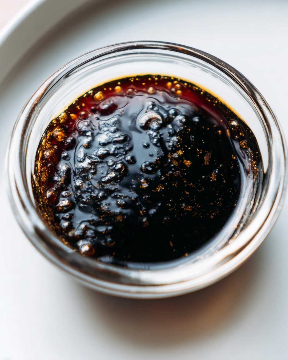 Close-up of thick, dark homemade teriyaki sauce with small bubbles and golden specks in a clear glass bowl.
