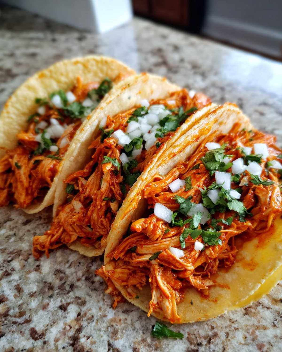 Three vibrant chicken tacos filled with shredded, saucy chicken and topped with diced white onion and fresh cilantro.