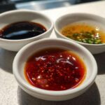 Three small white bowls containing different Dumpling Sauce recipes: chili oil, soy sauce, and a scallion vinaigrette.