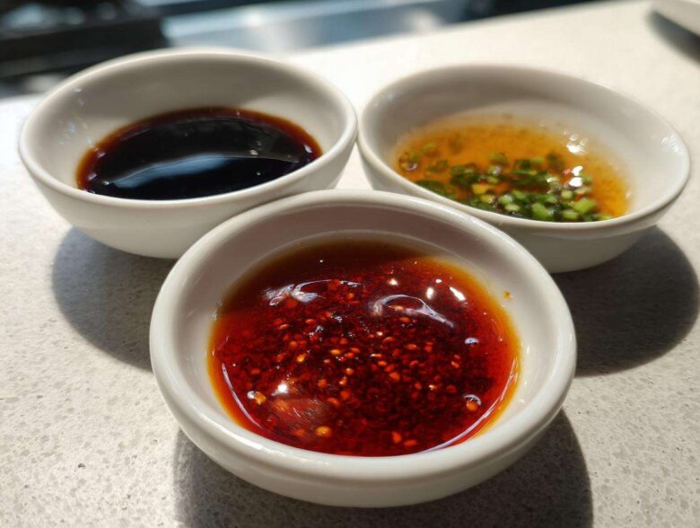 Three small white bowls containing different Dumpling Sauce recipes: chili oil, soy sauce, and a scallion vinaigrette.