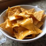 Close-up of tortilla chips generously covered in bright, smooth nacho cheese sauce in a white bowl.