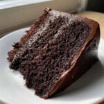 A close-up of a rich, dark slice of ultimate chocolate fudge cake with glossy frosting on a white plate.
