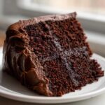 Close-up of a moist slice of ultimate chocolate fudge cake covered in thick chocolate frosting on a white plate.