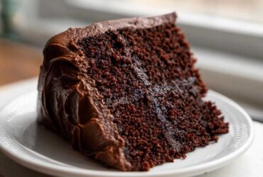 Close-up of a moist slice of ultimate chocolate fudge cake covered in thick chocolate frosting on a white plate.