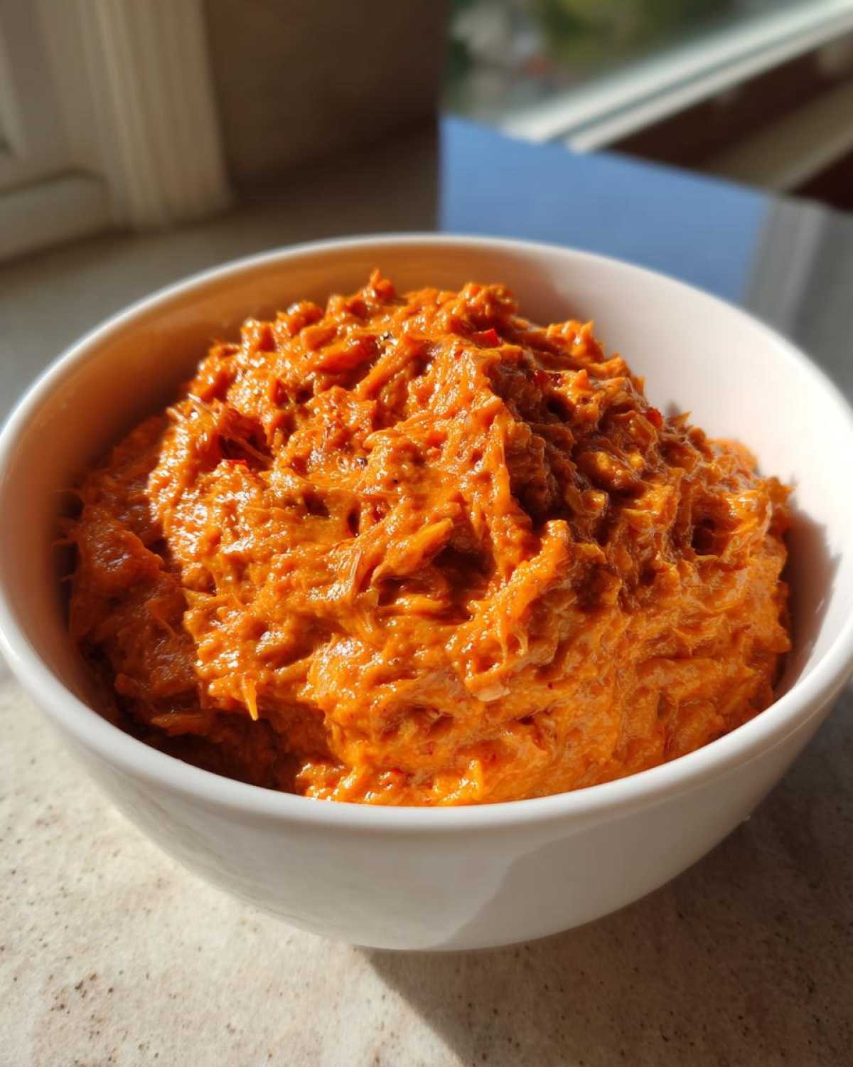 Close-up of vibrant orange chili cheese dip with a shredded texture served in a white bowl.