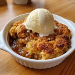 Close-up of a warm apple cobbler recipe served in a white ramekin, topped with melting vanilla ice cream.