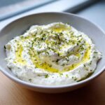 A close-up of a bowl filled with creamy whipped feta dip, drizzled with olive oil and topped with fresh herbs.