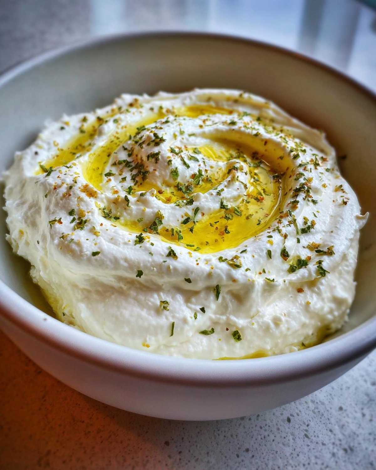 A close-up of a white bowl filled with creamy whipped feta dip, swirled with olive oil and sprinkled with herbs.