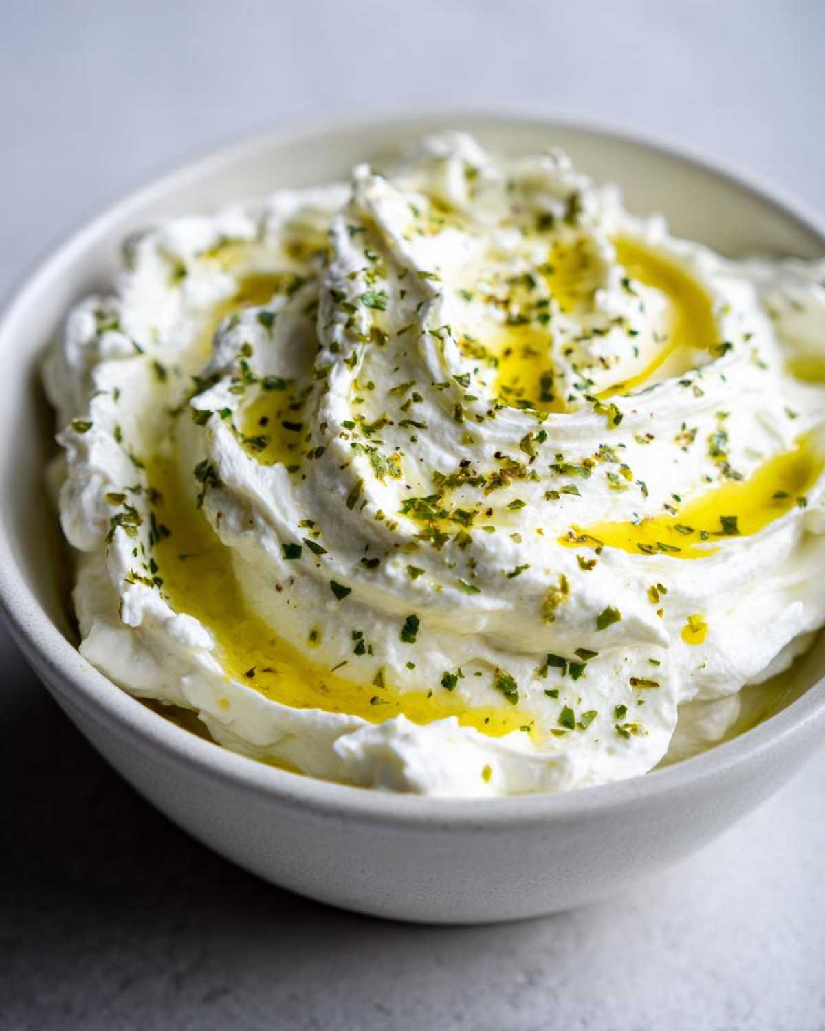 Close-up of creamy whipped feta dip swirled in a bowl, topped with olive oil and herbs.
