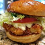 Close-up of a golden, crispy Air Fryer Crispy Fish Sandwich topped with melted sauce, tomato, and lettuce.