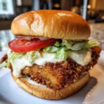 A close-up of an Air Fryer Crispy Fish Sandwich featuring a golden, crunchy fillet, lettuce, tomato, and generous tartar sauce on a brioche bun.