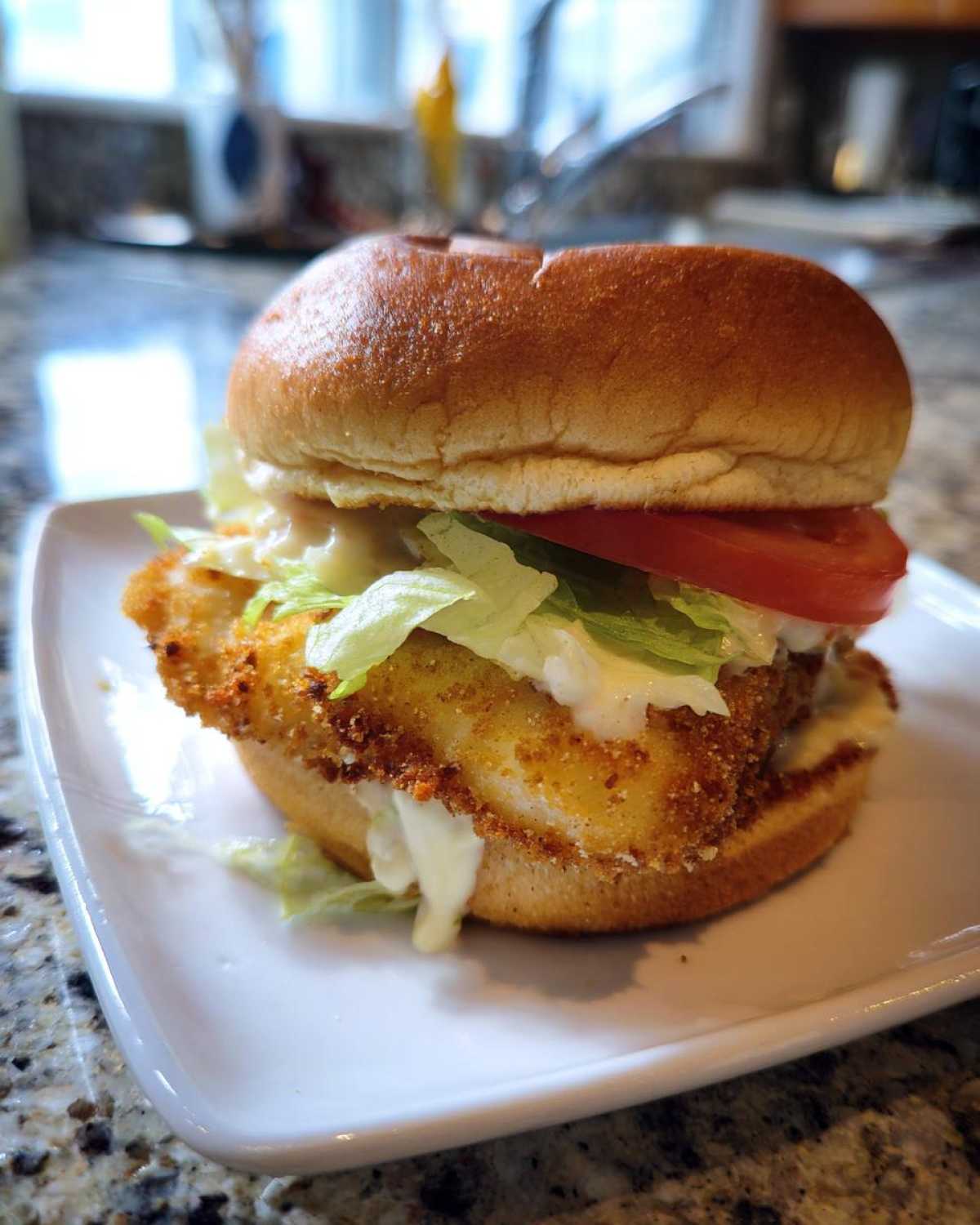 A close-up of an Air Fryer Crispy Fish Sandwich featuring a golden, breaded fillet, lettuce, tomato, and tartar sauce on a toasted bun.