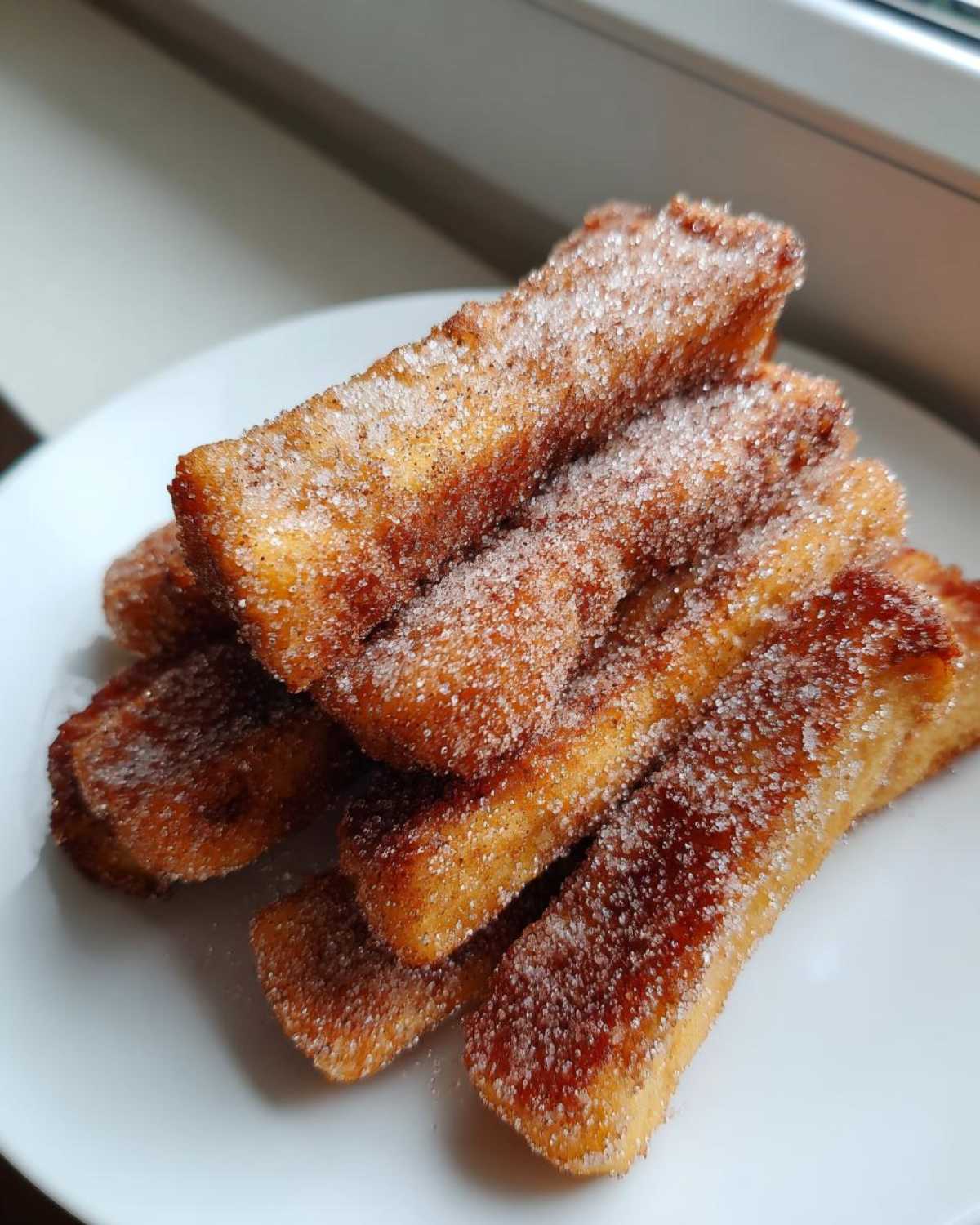 A pile of golden brown Air Fryer French Toast Sticks heavily coated in sparkling cinnamon sugar on a white plate.