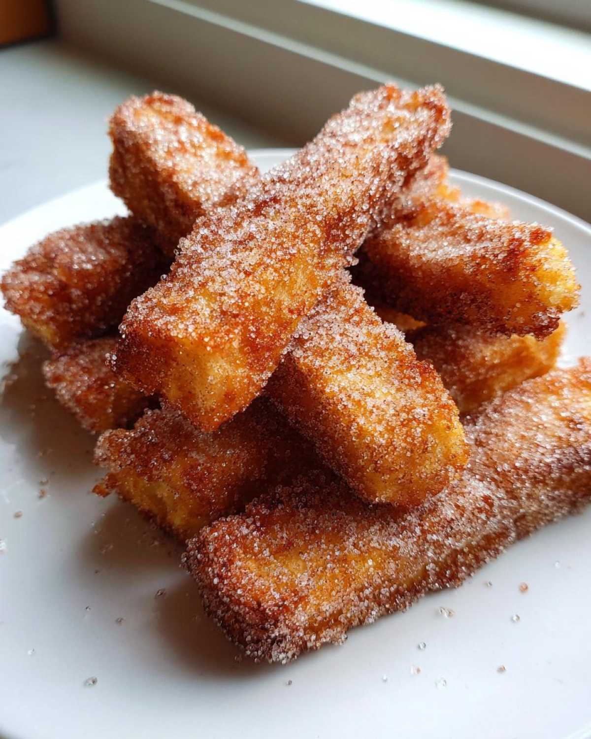 A pile of golden brown Air Fryer French Toast Sticks heavily coated in sparkling cinnamon sugar on a white plate.