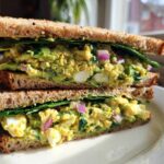 A close-up of an Avocado Egg Salad Sandwich cut in half, showing creamy filling, egg chunks, red onion, and spinach on whole wheat bread.
