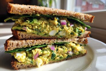 A close-up of an Avocado Egg Salad Sandwich cut in half, showing creamy filling, egg chunks, red onion, and spinach on whole wheat bread.