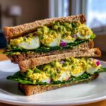 A tall, stacked Avocado Egg Salad Sandwich featuring sliced hard-boiled eggs, mashed avocado, and red onion on whole wheat bread.