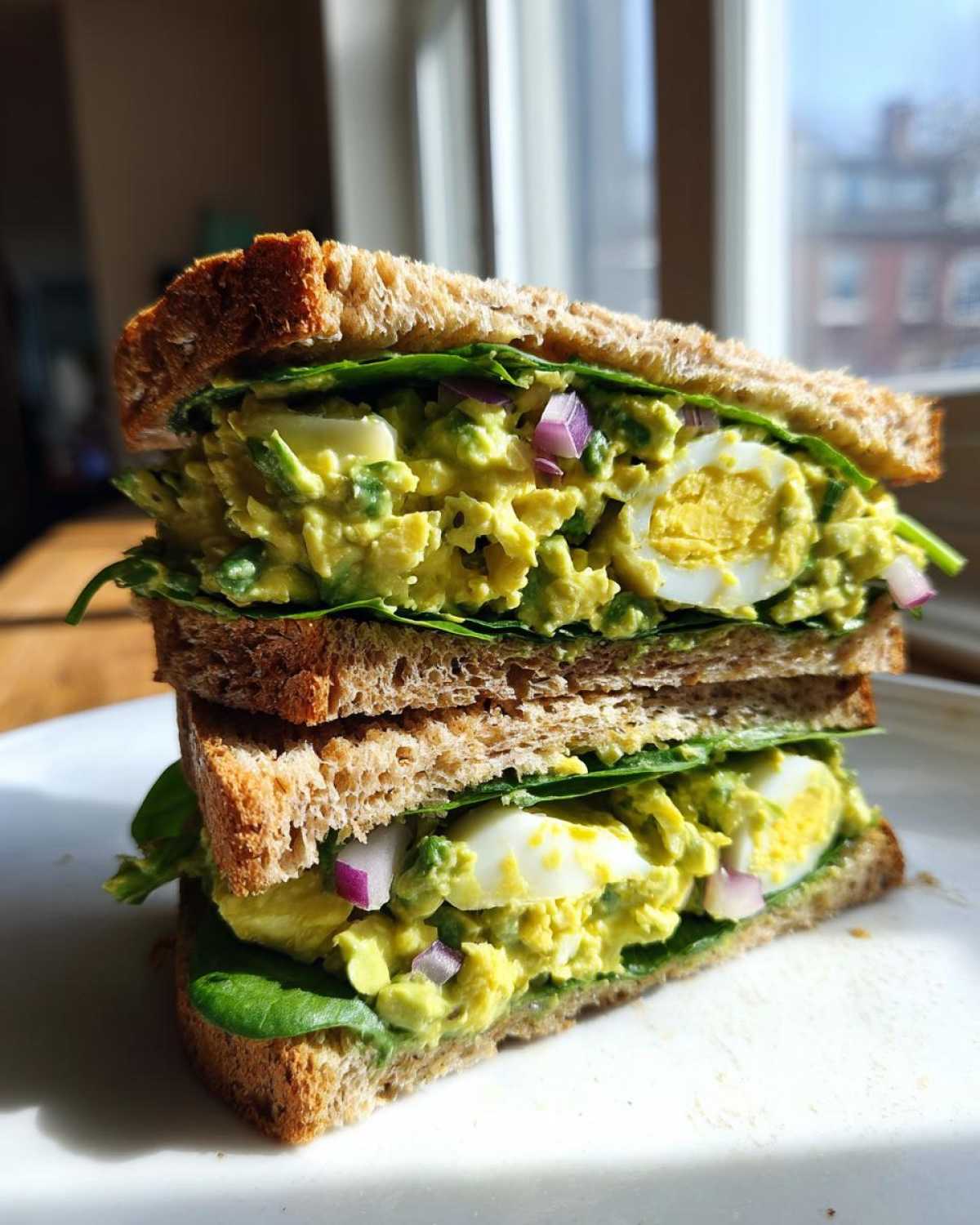 A tall, stacked Avocado Egg Salad Sandwich featuring creamy filling, sliced hard-boiled eggs, red onion, and spinach on whole wheat bread.