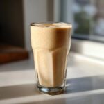 A tall glass filled with a thick, frothy Banana Peanut Butter Smoothie, catching morning sunlight.