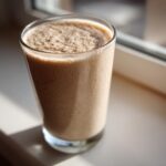 A tall glass filled with a creamy, frothy Banana Peanut Butter Smoothie sitting on a sunlit windowsill.