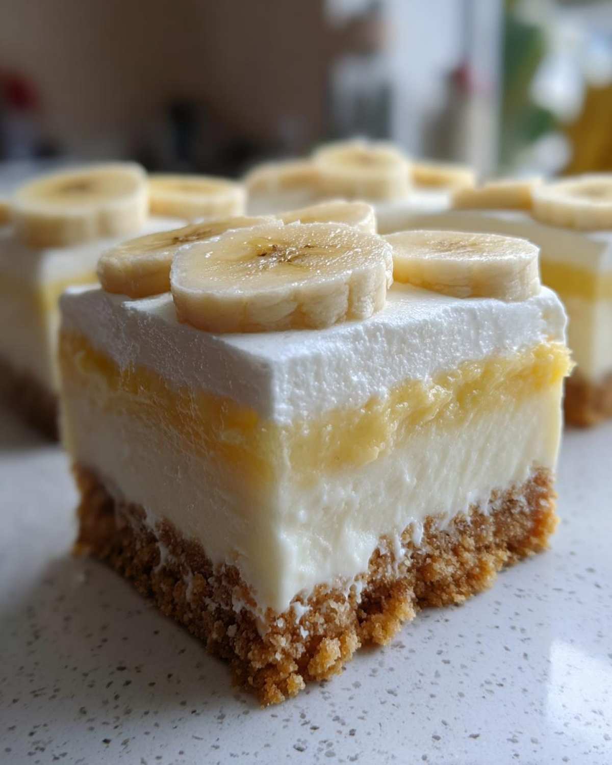 A close-up of a layered Banana Pudding Cheesecake Bars slice showing graham cracker crust, creamy filling, and banana topping.