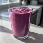 A tall glass filled with a thick, vibrant purple Blueberry Lemon Chia Smoothie sitting on a sunlit countertop.