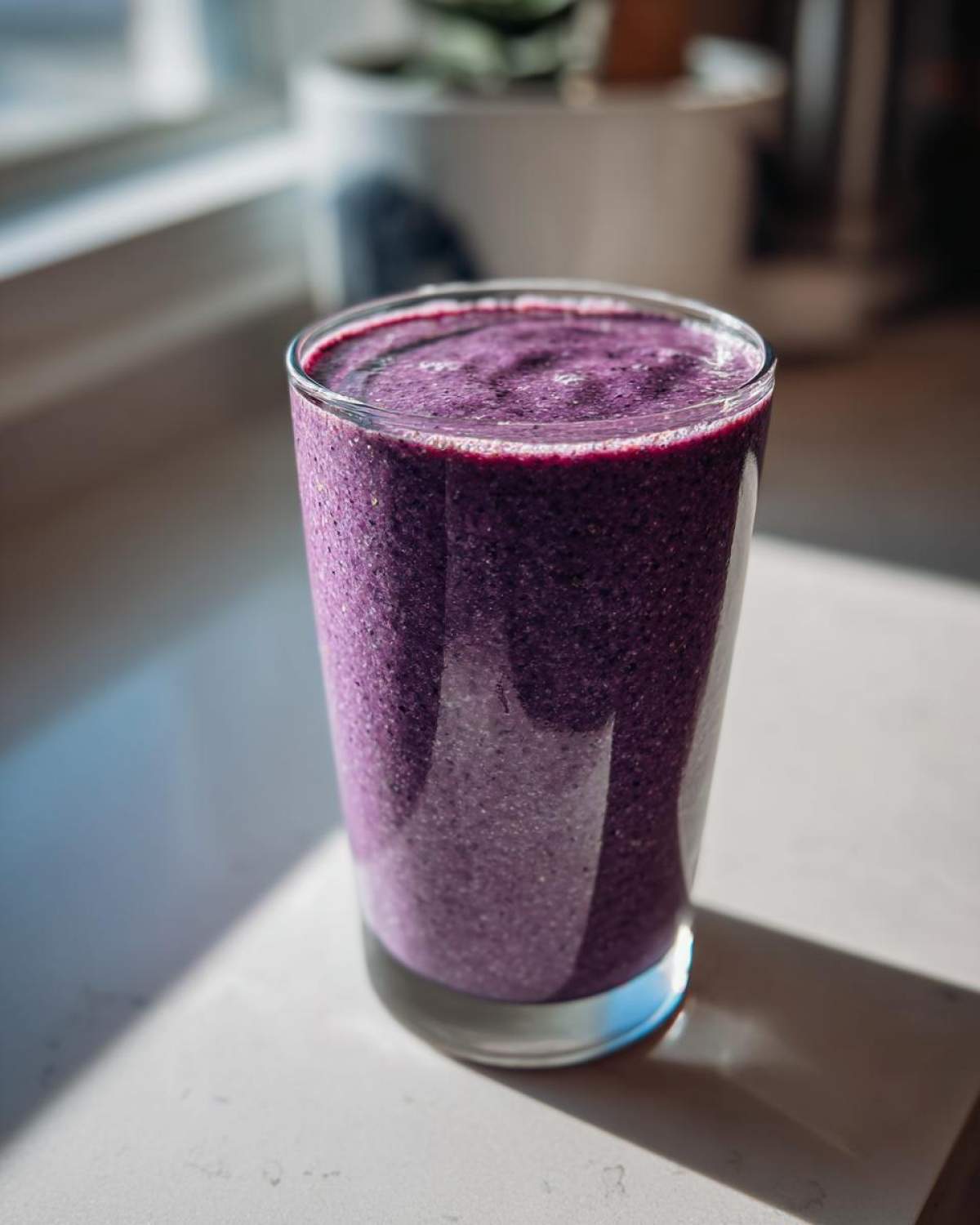 A tall glass filled with a thick, vibrant purple Blueberry Lemon Chia Smoothie sitting on a light countertop.
