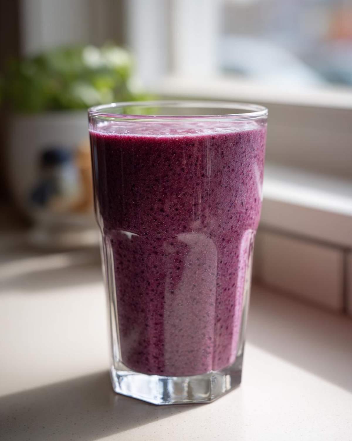 A tall glass filled with a thick, deep purple Blueberry Lemon Chia Smoothie sitting on a light countertop near a window.