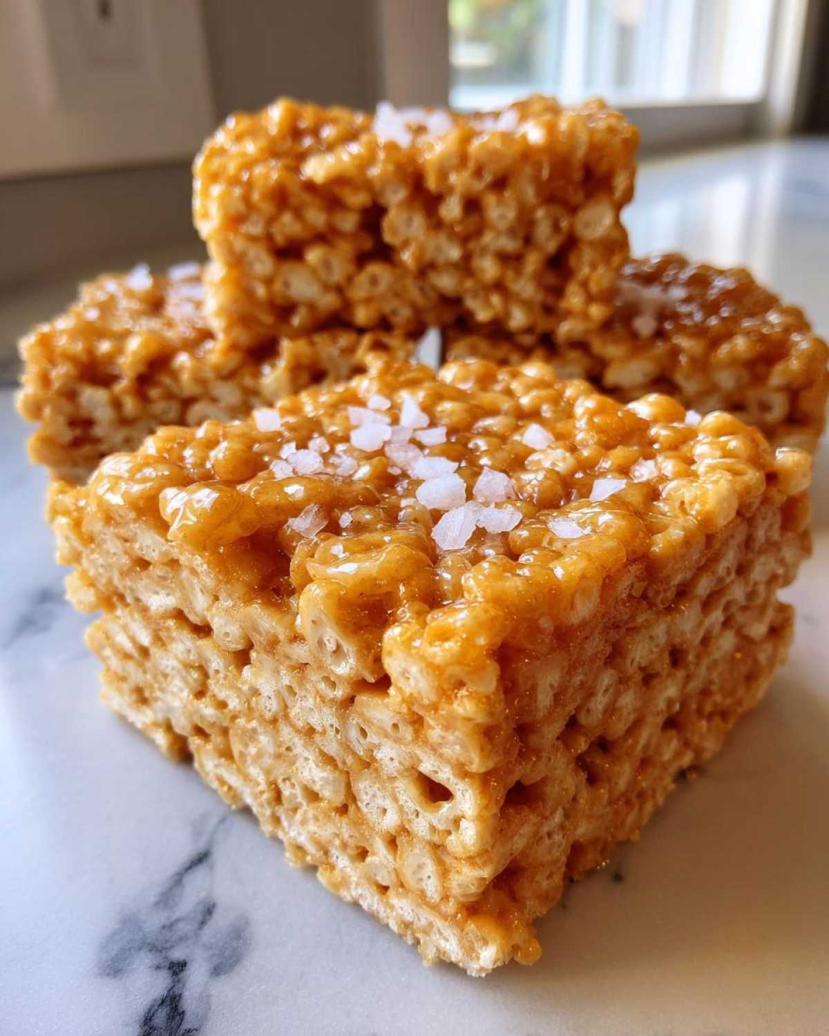 A close-up of thick Brown Butter Rice Krispie Treat Bars topped with flaky sea salt on a marble surface.