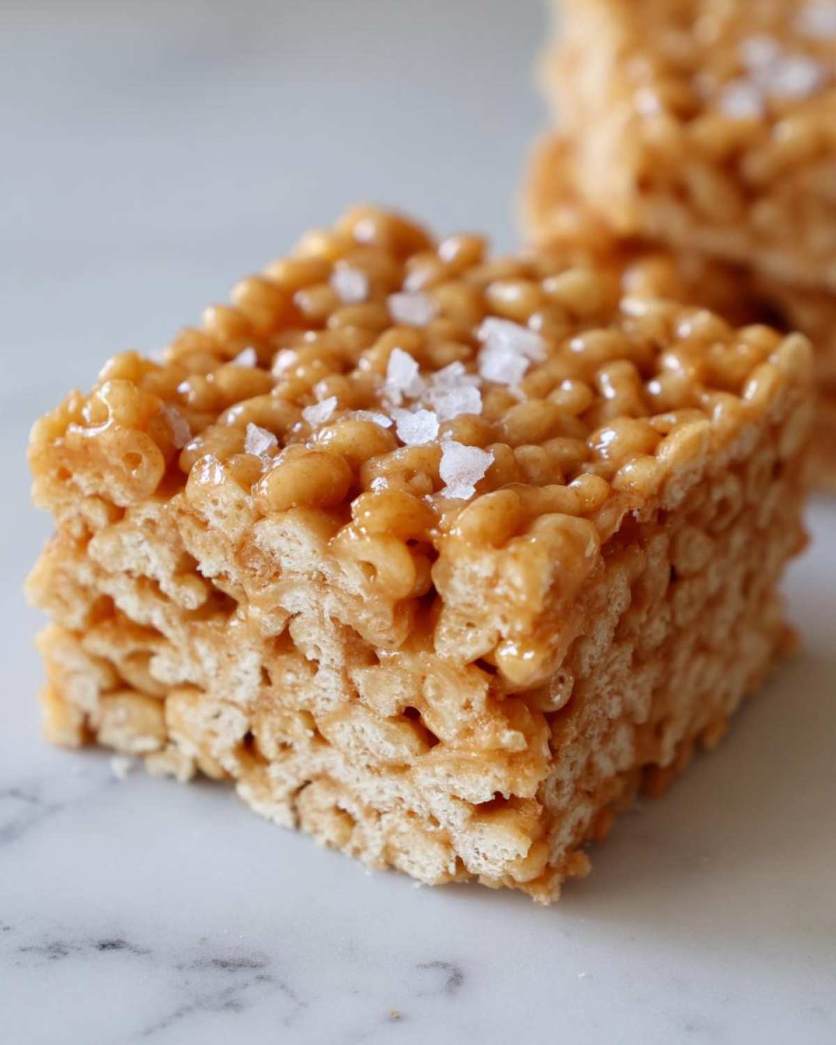 A close-up of a square Brown Butter Rice Krispie Treat Bar topped with flaky sea salt, resting on a white marble surface.