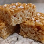 Three stacked Brown Butter Rice Krispie Treat Bars topped with flaky sea salt on a granite counter.