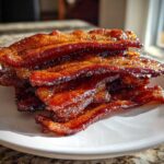 A close-up stack of crispy, shiny Brown Sugar Glazed Bacon on a white plate.