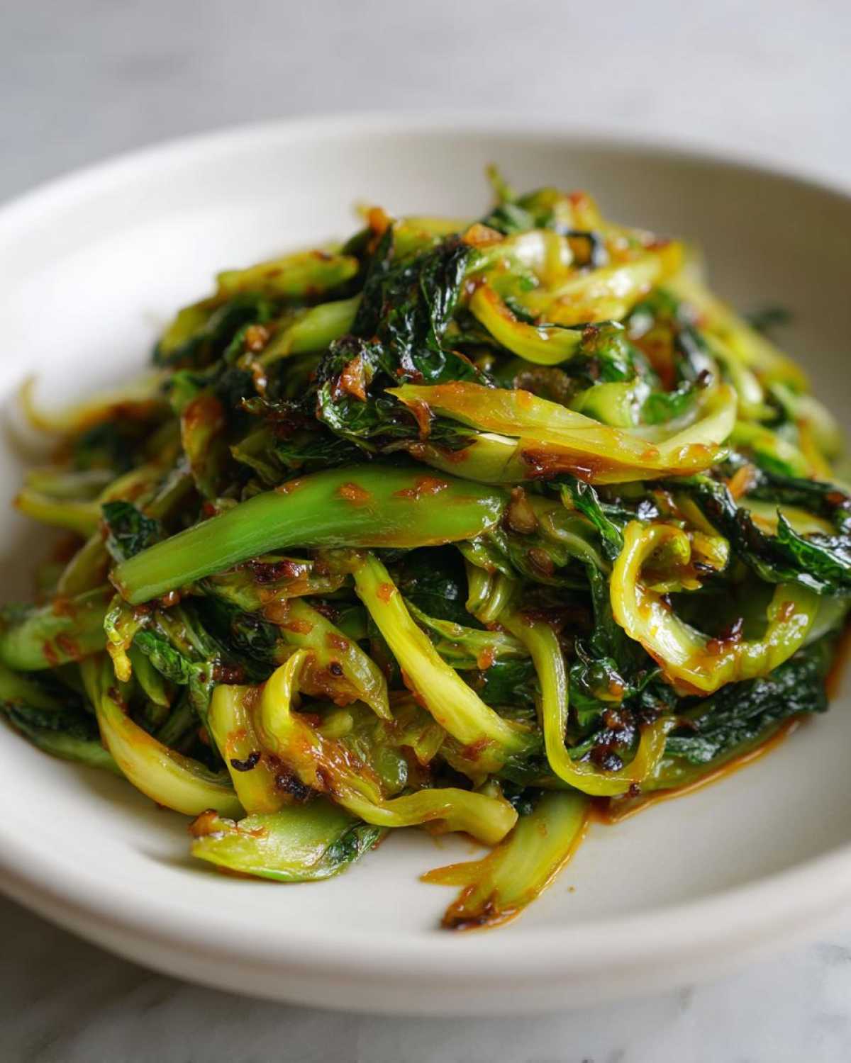 A close-up shot of vibrant green, wilted cabbage stir fry coated in a glossy garlic ginger sauce, served in a white bowl.