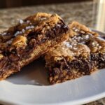 Two rich, chewy chocolate chip cookie bars cut and stacked slightly, showing melted chocolate centers and sprinkled with flaky sea salt.