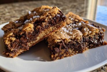 Two rich, chewy chocolate chip cookie bars cut and stacked slightly, showing melted chocolate centers and sprinkled with flaky sea salt.
