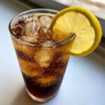 Close-up of a tall glass filled with bubbly dark liquid, ice cubes, and garnished with a bright lemon slice, representing Cold Brew Lemonade.