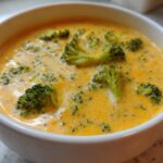 Close-up of a white bowl filled with creamy, orange-yellow Copycat Broccoli Cheddar Soup topped with bright green broccoli florets.