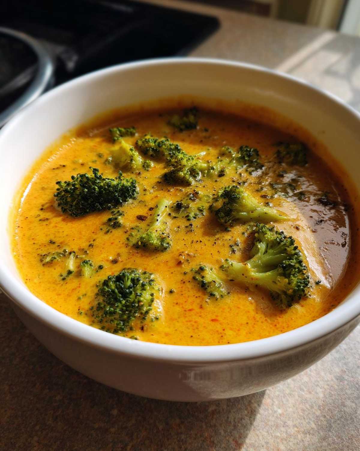 Close-up of a white bowl filled with creamy, orange-hued Copycat Broccoli Cheddar Soup topped with bright green broccoli florets.