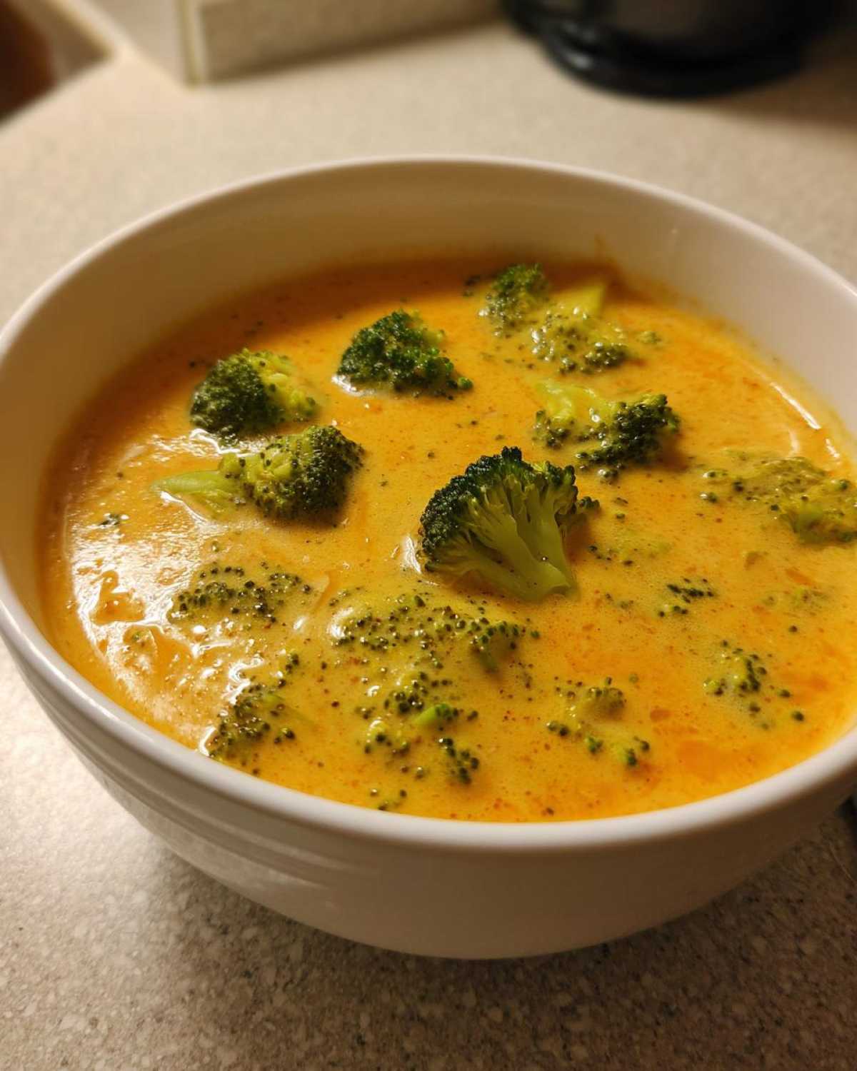 Close-up of a white bowl filled with thick, orange-yellow Copycat Broccoli Cheddar Soup topped with florets.