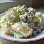A close-up of a serving of Creamy Dill Potato Salad mixed with red onion and fresh dill on a white plate.