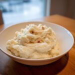 A white bowl filled with fluffy Creamy Garlic Mashed Cauliflower, garnished with small brown bits, sitting on a wooden table.