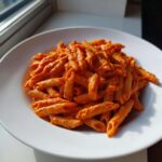 A white bowl filled with penne pasta coated in vibrant orange Creamy Roasted Red Pepper Pasta sauce.