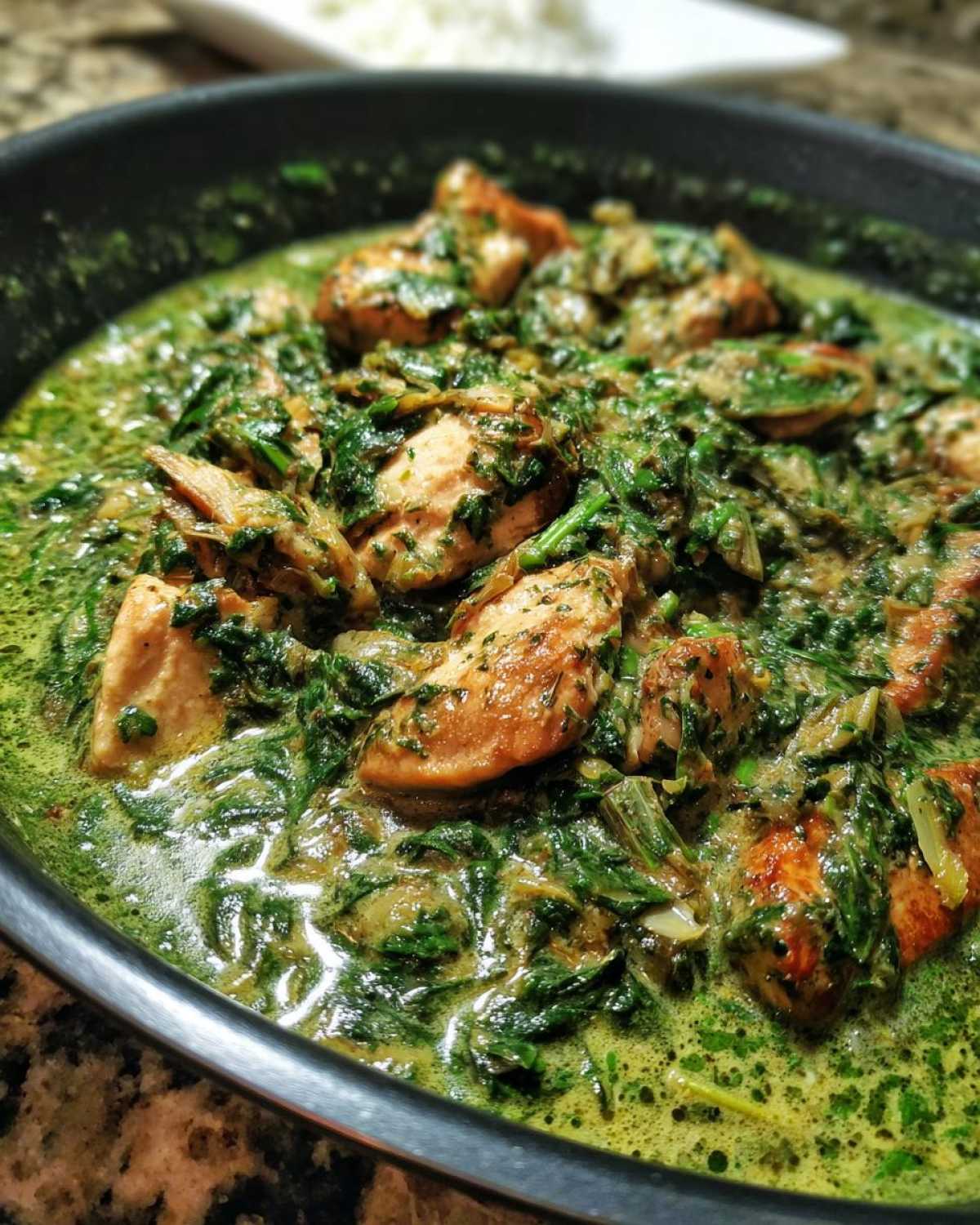 Close-up view of seared chicken pieces simmering in a rich, green, creamy spinach artichoke sauce in a black skillet.