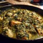 Close-up of seared chicken pieces simmering in a rich, creamy spinach artichoke sauce in a black skillet.